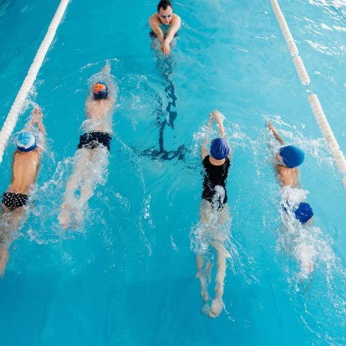 A group of boys and girls train and learn to swim in a modern swimming pool with an instructor. Development of children's sports. Healthy parenting and promotion of children's sports.